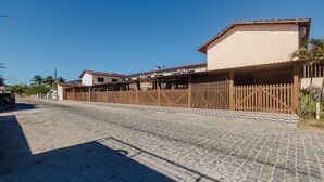 A street with buildings, a wooden fence, and a clear blue sky. - Cond. c\ Piscina e Churrasqueira a 650 m Praia de Taperapuan \ PFNs (Porto Seguro)