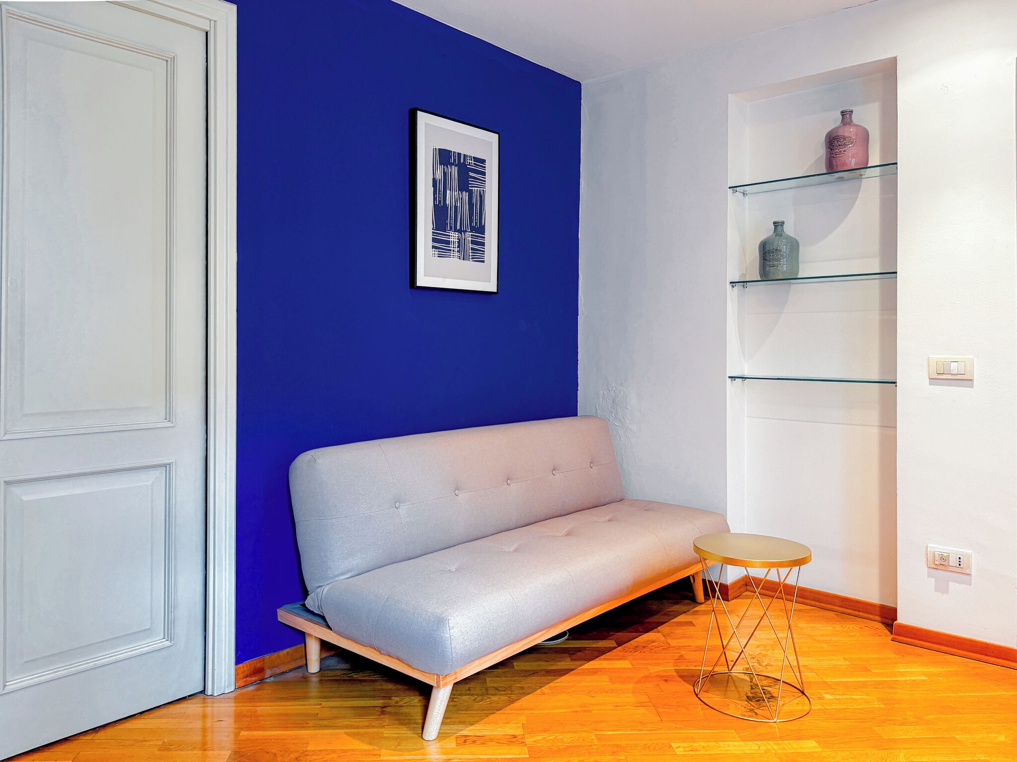 Ein Zimmer mit blauer Wand, einem grauen Sofa, einem kleinen runden Tisch und einem Glastischchen mit Flaschen.