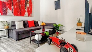 Una sala de estar moderna con un sofá gris, un cuadro abstracto rojo y blanco, un pequeño coche de juguete rojo y una planta en maceta.