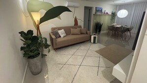 Ein modernes Wohnzimmer mit einem beigefarbenen Sofa, einer großen grünen Wanddekoration, einer Essecke mit rundem Tisch und Stühlen sowie einer Deckenleuchte.