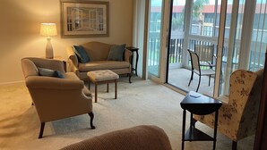 Un salon comprenant un canapé, un fauteuil, un pouf et une table. On y trouve une lampe, un tableau encadré accroché au mur, ainsi qu’une porte-fenêtre coulissante donnant sur un espace ext érieur avec une table et des chaises de patio.