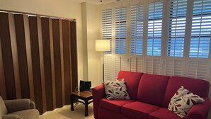 Un salon avec un canapé rouge, un fauteuil beige, une petite table avec une lampe et une fenêtre avec des stores.