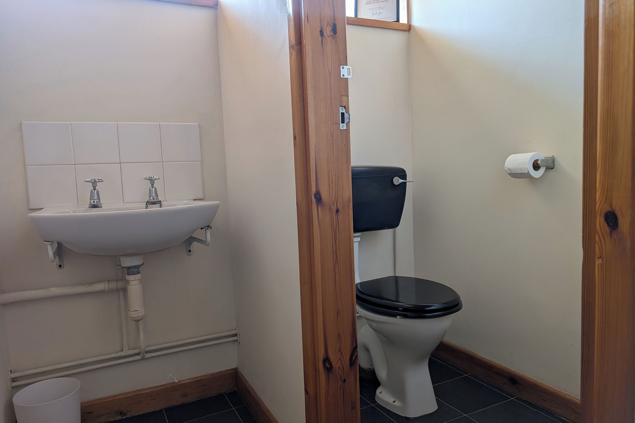 A bathroom with a white sink, two faucets, a toilet with a black seat, and a roll of toilet paper on the wall.