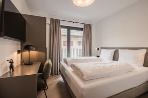 A modern hotel room with a bed, a desk, a chair, a lamp, and a view of buildings through the window. - Apartment Near Schaterzlift Kirchberg (Kirchberg in Tirol)