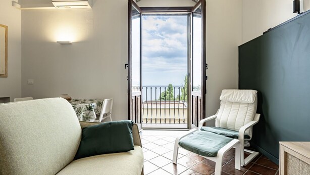 Ein Zimmer mit Bett, Stuhl, Tisch, einem Balkon mit Geländer und einer Tür zum Außenbereich.