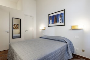 A bedroom with a bed, a door, a framed picture of lighthouses, and a wall-mounted light fixture.