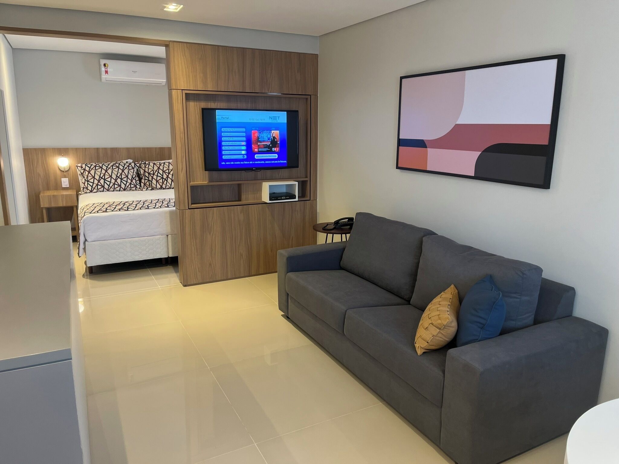 Uma sala de estar moderna com um sofá cinzento, uma TV presa a um painel de madeira e uma cama com almofadas estampadas ao fundo.
