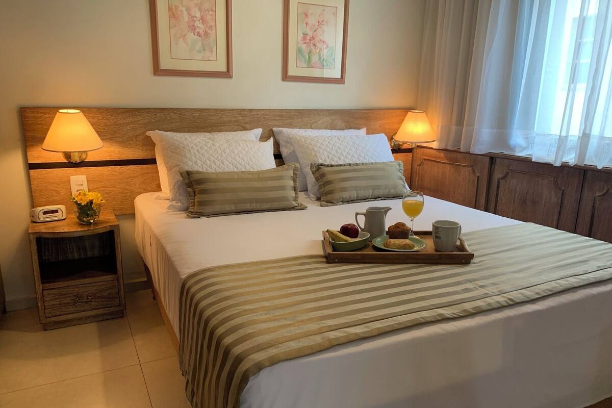 Una cama bien hecha con una colcha a rayas, dos almohadas, una mesita de noche con una lámpara, una bandeja con productos de desayuno y un cuadro con motivos florales en la pared.