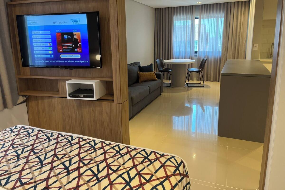 Una habitación de hotel con televisión montada en la pared, un sofá, una mesa de comedor con sillas y un armario pequeño.