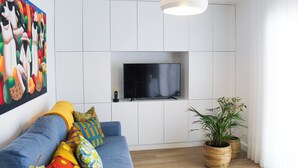 Una sala de estar moderna con un sofá azul, cojines coloridos, un televisor de pantalla plana montado en una pared blanca y dos grandes macetas tejidas.