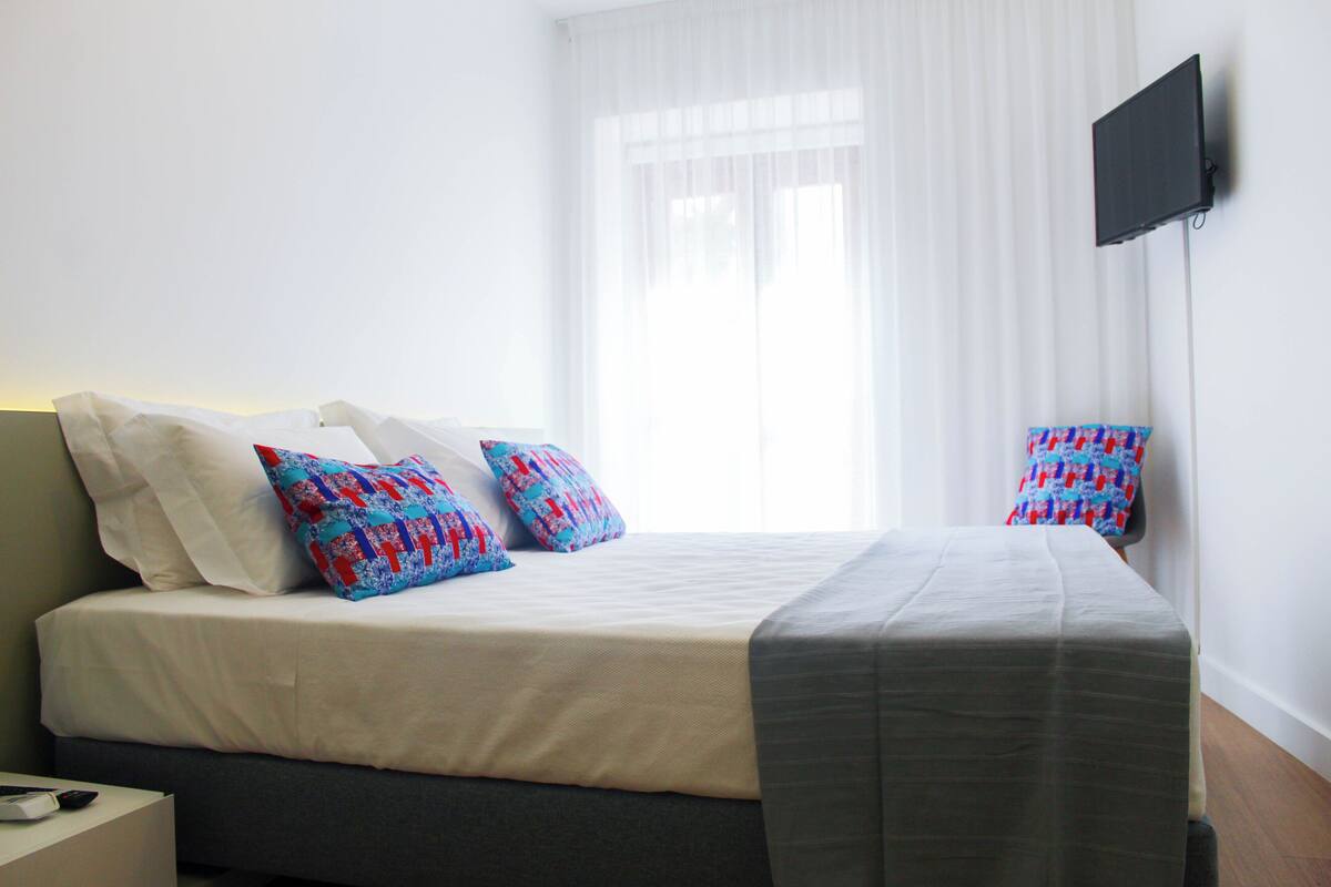 Ein ordentlich bezogenes Bett mit Kissen in Weiß und Blau, einer grauen Decke und einem flachbildfernseher an der Wand.