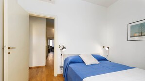 A bedroom with a bed, bedside lamps, a door leading to another room, and a framed picture on the wall.