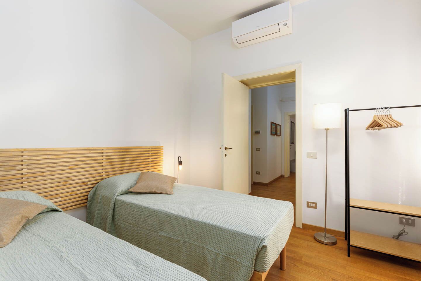 A bedroom with two beds, a wooden headboard, a bedside lamp, an air conditioning unit, and a wardrobe with a hanging rack.