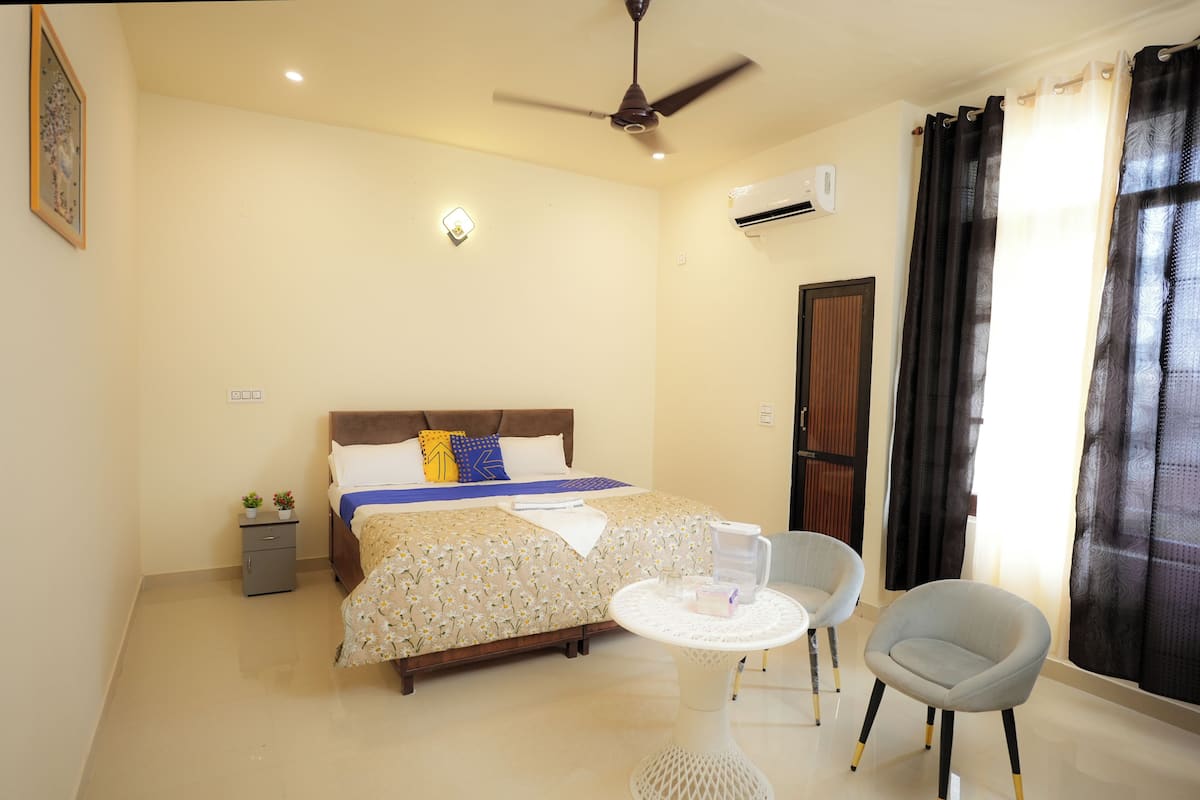 A hotel room with a bed, bedside table, two chairs, a ceiling fan, and a window with curtains.