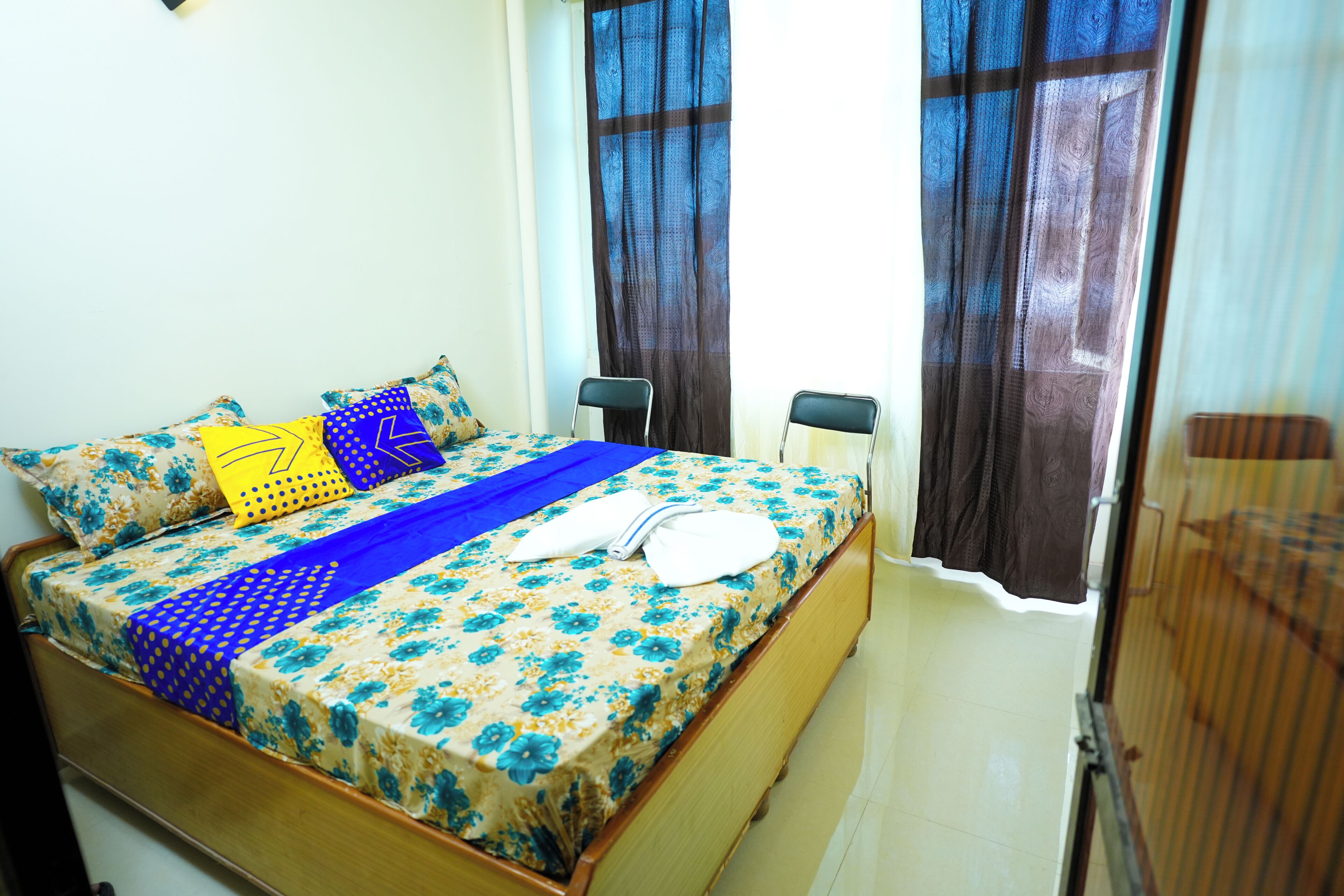 A neatly made bed with floral bedding, a blue blanket, and a white towel. There are two chairs and a small table in the room.