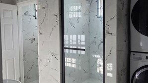 A bathroom with white marble tiles, a glass shower enclosure, a white washing machine, and a striped rug. - 3 min from Ogle Intl Airport 
Vacation Home in Prime Location (Ogle)