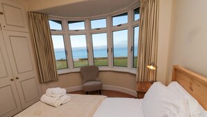 A hotel room with a large bed, a chair, and a view of the sea through multiple windows. - Glenavon (Seahouses)
