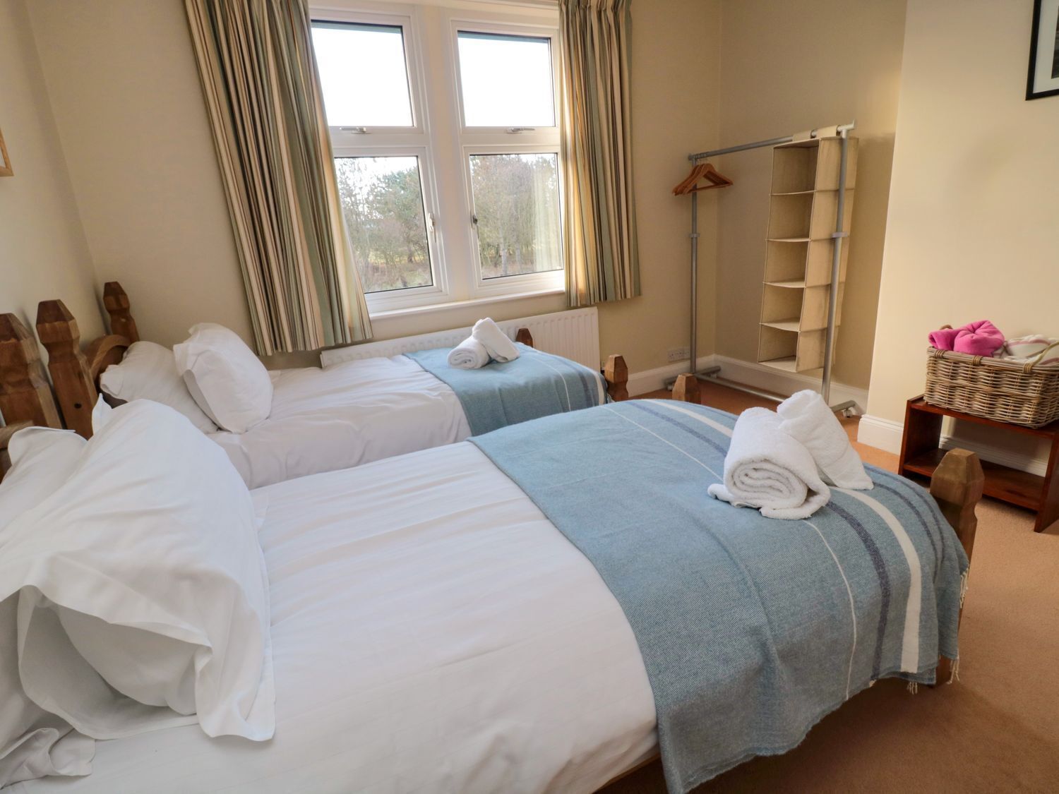 A hotel room with two beds, a window with curtains, a wooden headboard, a radiator, a wooden shelf, and a basket with pink items.