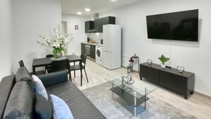 A modern living room with a sofa, a glass coffee table, and a dining area with chairs. There is a kitchen with a washing machine and a TV mounted on the wall. - Naffah Apartments II (Madrid)