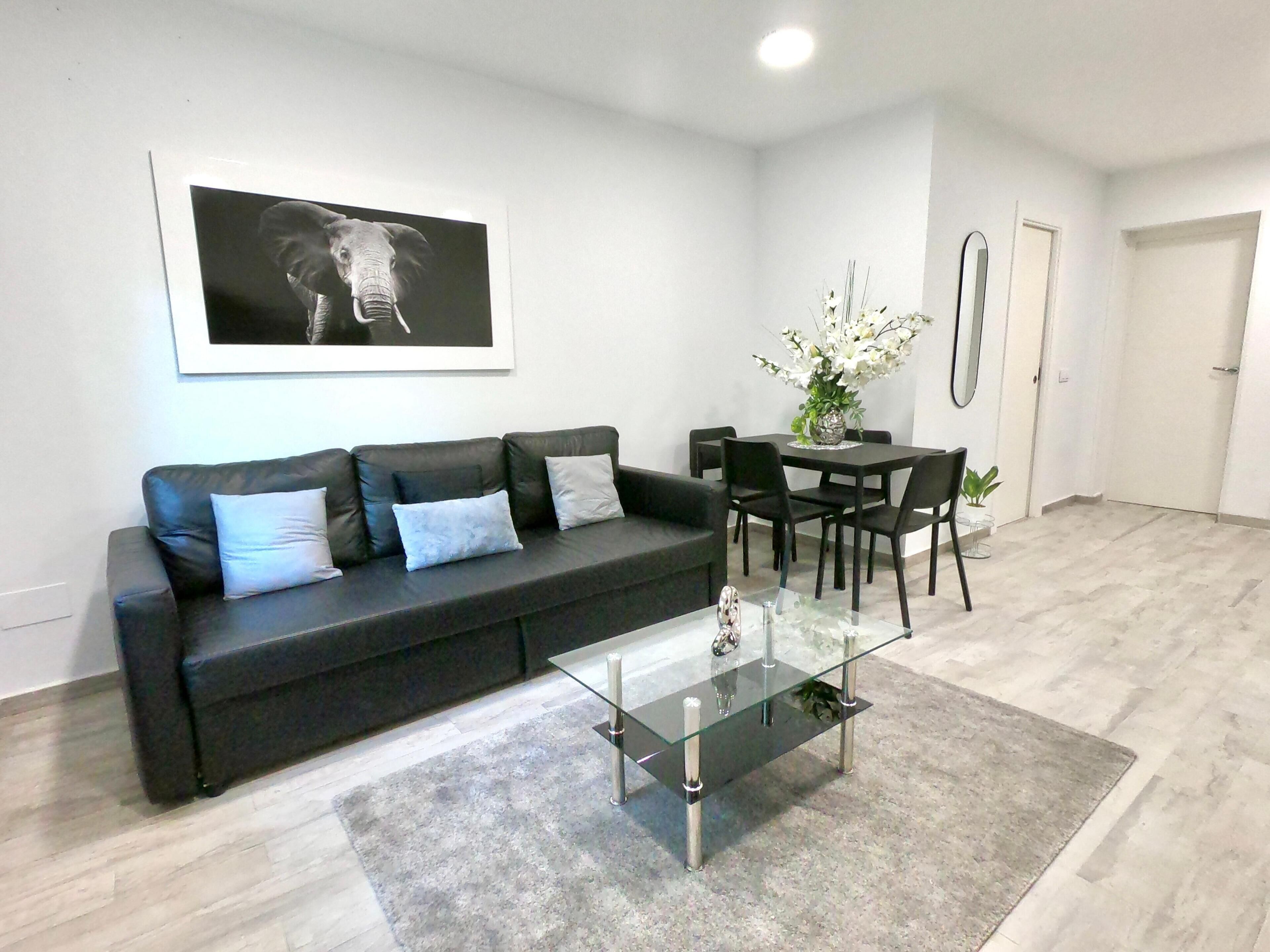 A modern living room with a black leather sofa, a glass coffee table, and a dining area with a white table and chairs.