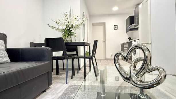 A modern living room with a sofa, a dining table with chairs, a decorative heart-shaped sculpture, and a kitchen area with cabinets and a microwave. - Naffah Apartments II (Madrid)