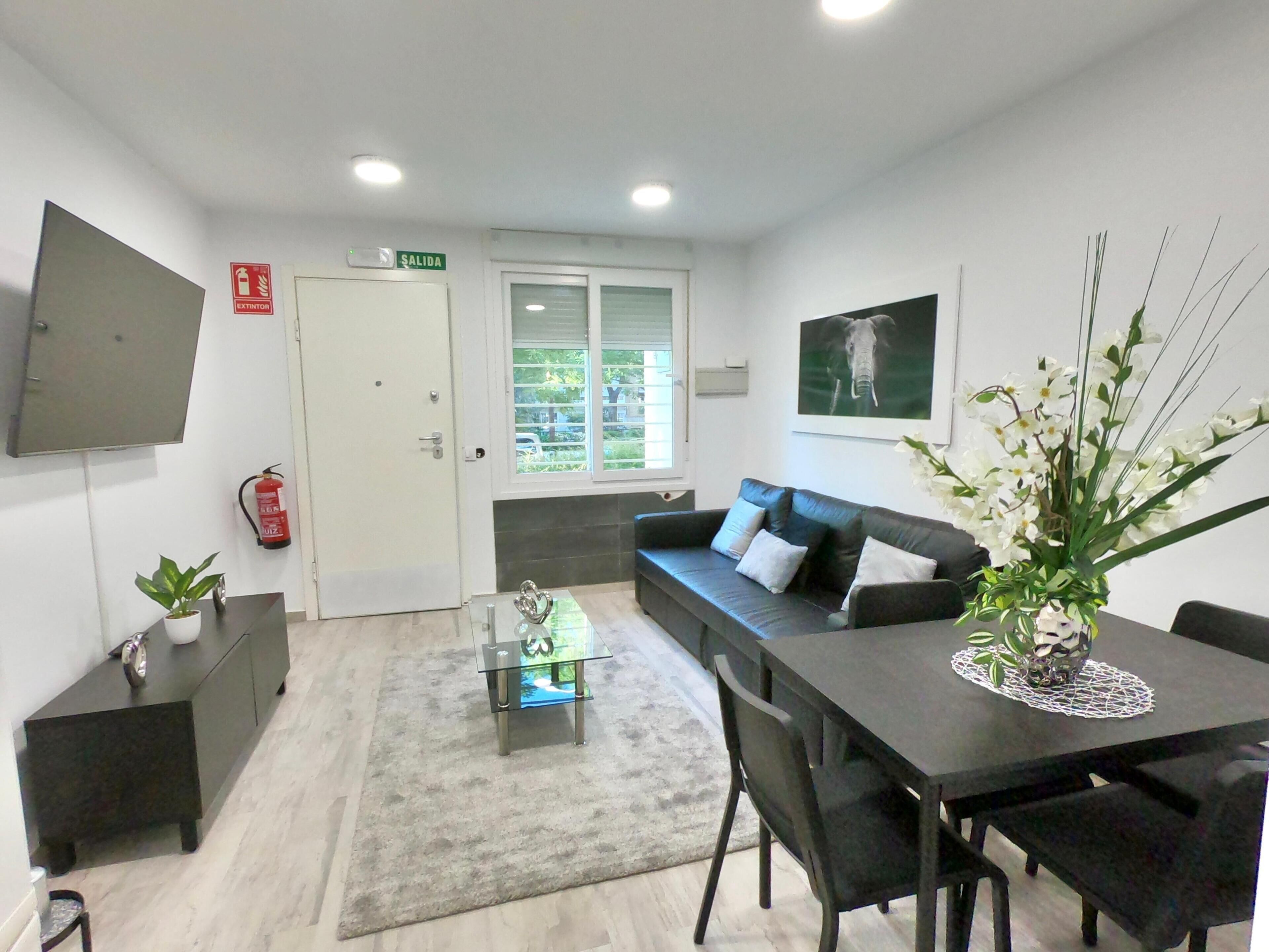 Una sala de estar moderna con un sofá, una mesa de centro y una mesa de comedor. Hay un extintor de incendios montado en la pared y una puerta con un letrero que indica una salida.
