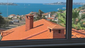 Vista costeira pela janela, com uma cidade de edifícios com telhados vermelhos, mar azul límpido e uma ilha ao longe.