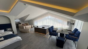 Quarto de hotel moderno com uma cama grande, um sofá, área de jantar com cadeiras, uma mesa pequena, hidromassagem e uma janela grande com cortinas.