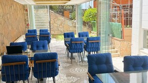 Uma área de refeições com cadeiras estofadas em azul e mesas, piso de azulejos estampados e vista para um jardim através de portas de vidro.