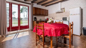 A dining area with a red tablecloth, wooden chairs, a kitchen with a white refrigerator, and a view through a glass door. - Non-air-conditioned country cottage near a farm (Questembert)
