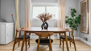 Un comedor con una mesa y sillas de madera, un florero con flores secas, una planta en maceta y una obra de arte en la pared.