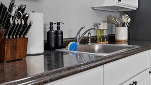 Una cocina con encimera de granito, un juego de cuchillos, toallas de papel, jabón de manos, una botella de detergente para platos, una esponja azul y un lavabo de acero inoxidable.