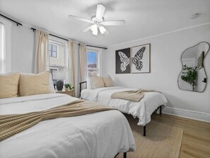 A bedroom with two beds, a ceiling fan, a window with curtains, and a wall-mounted mirror.