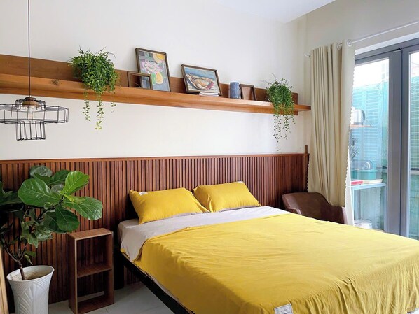 Ein Schlafzimmer mit einem Bett, gelber Bettwäsche, einem Holz-Kopfende, einer hängenden Pflanze, einem Regal mit Bildern und einem Fenster mit Vorhängen.