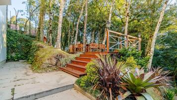 Una escalera de madera conduce a un espacio cubierto con asientos, rodeado de abundante follaje y árboles.