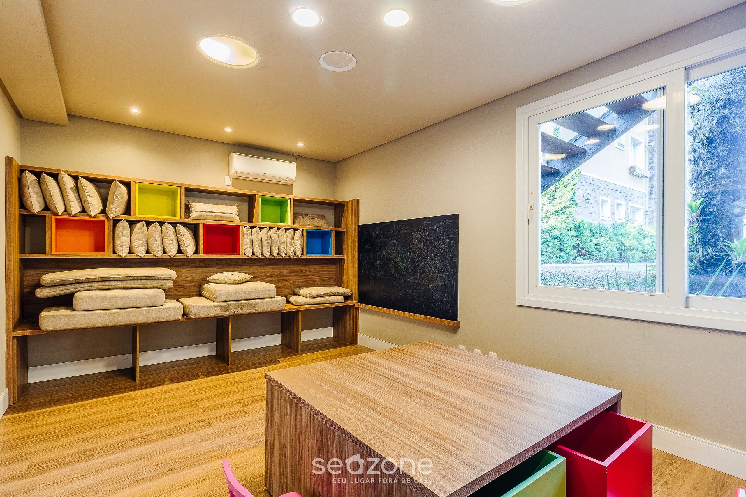 Una habitación moderna con suelo de madera, una mesa de madera y una ventana con vistas a zonas verdes. Se observan estanterías con almohadas y cajas de almacenamiento.