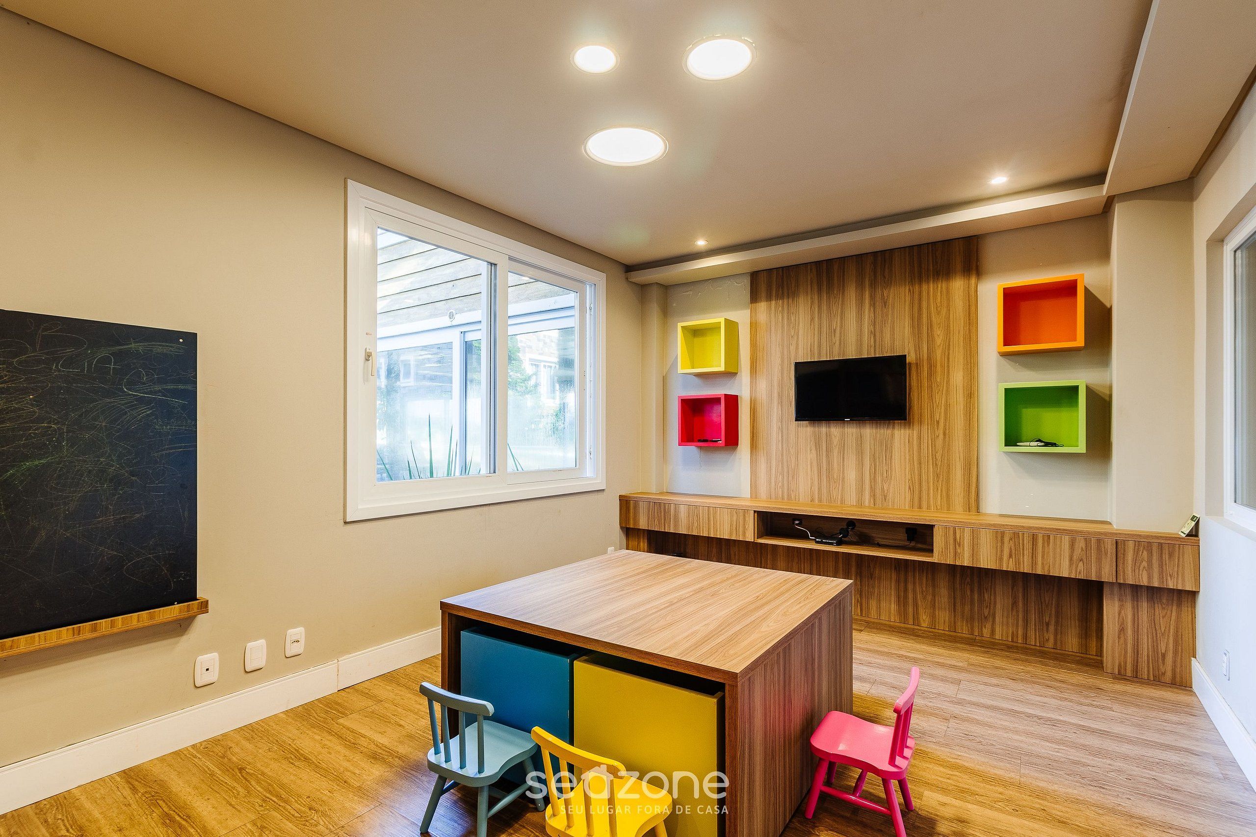 A modern children's playroom with a wooden table, colorful chairs, a chalkboard, and built-in shelves.