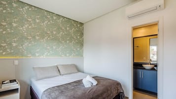 Un dormitorio con cama, almohadas, una mesita de noche con libros, una pared con papel tapiz floral y un baño visible a través de una puerta abierta.