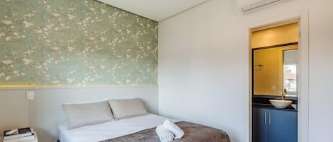 A bedroom with a bed, pillows, a bedside table with books, a wall with floral wallpaper, and a bathroom visible through an open door.