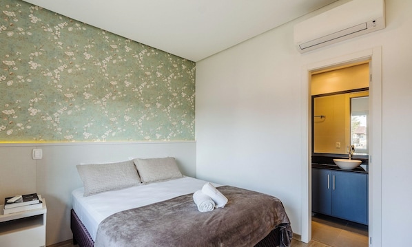 A bedroom with a bed, pillows, a bedside table with books, a wall with floral wallpaper, and a bathroom visible through an open door. - Beautiful Apartment With Heated Pool Cap0302 (Gramado)
