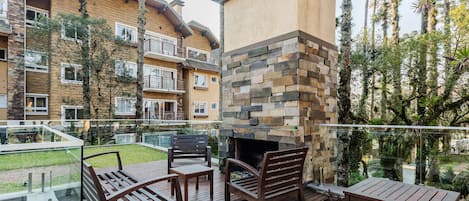 A wooden deck with outdoor furniture, a stone fireplace, and a view of a multi-story building with balconies.