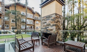 A wooden deck with outdoor furniture, a stone fireplace, and a view of a multi-story building with balconies. - Beautiful Apartment With Heated Pool Cap0302 (Gramado)