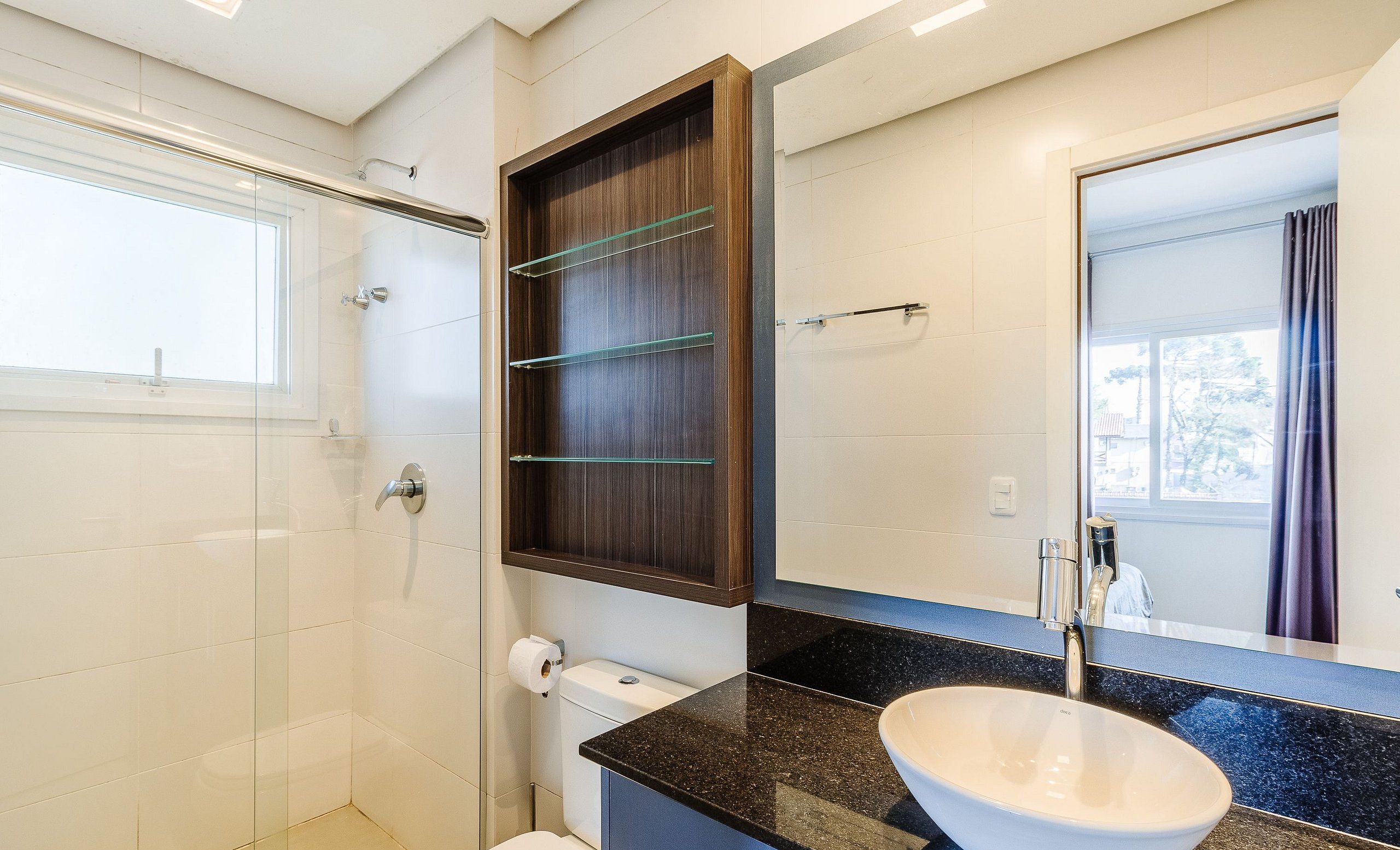 Un baño moderno con mampara de vidrio, un mueble de baño de madera oscura, encimera de granito negro y lavabo blanco.