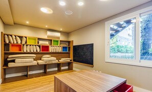 A modern room with a wooden floor, a wooden table, and a window with a view of greenery. Shelves with pillows and storage boxes are visible. - Beautiful Apartment With Heated Pool Cap0302 (Gramado)