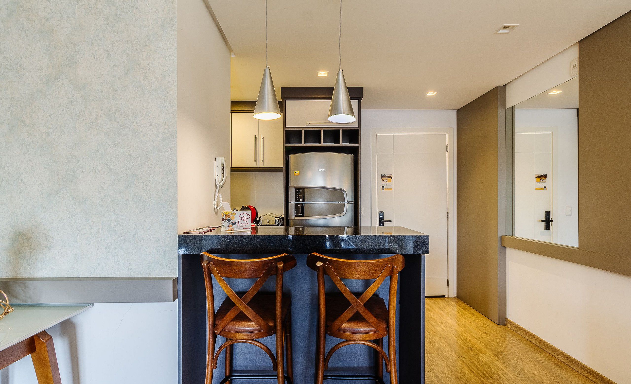 Una cocina moderna con una isla central, dos taburetes de madera, iluminación colgante y un refrigerador de acero inoxidable.