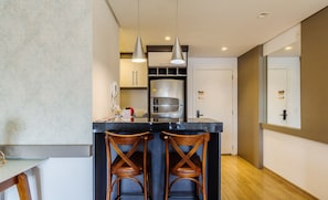 A modern kitchen with a central island, two wooden bar stools, pendant lighting, and a stainless steel refrigerator. - Beautiful Apartment With Heated Pool Cap0302 (Gramado)