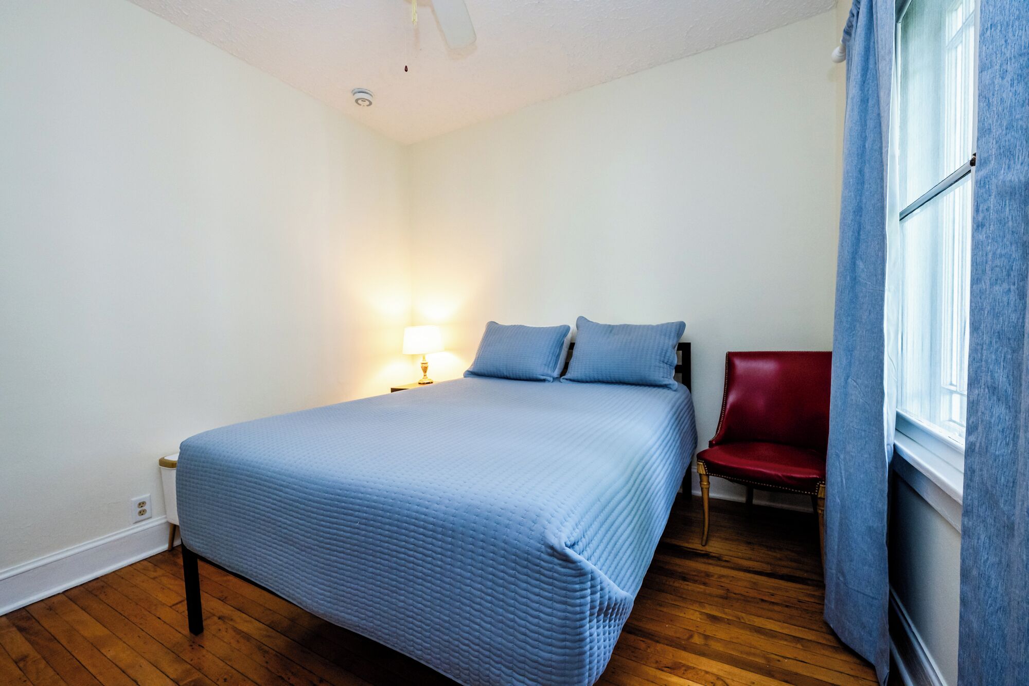 Ein Schlafzimmer mit einem Bett, einem Nachttisch mit Lampe, einem roten Sessel und einem Fenster mit Vorhängen.