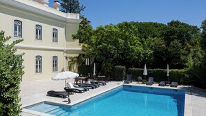 A pool area with lounge chairs and umbrellas, surrounded by greenery and a building with multiple windows. - Royal Hideaway – Estoril 2BR Pool Retreat (Cascais)