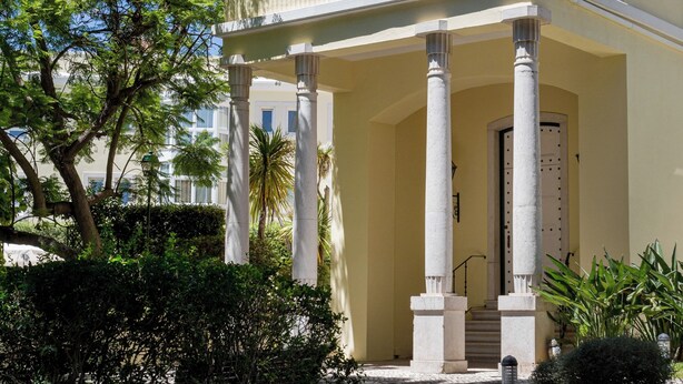 A two-story building with a white facade, large columns supporting a balcony, and a clear blue sky in the background. - Royal Hideaway – Estoril 2BR Pool Retreat (Cascais)