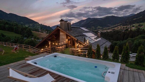 Een houten terras met een jacuzzi en ligstoelen, met uitzicht op een bergachtig landschap.